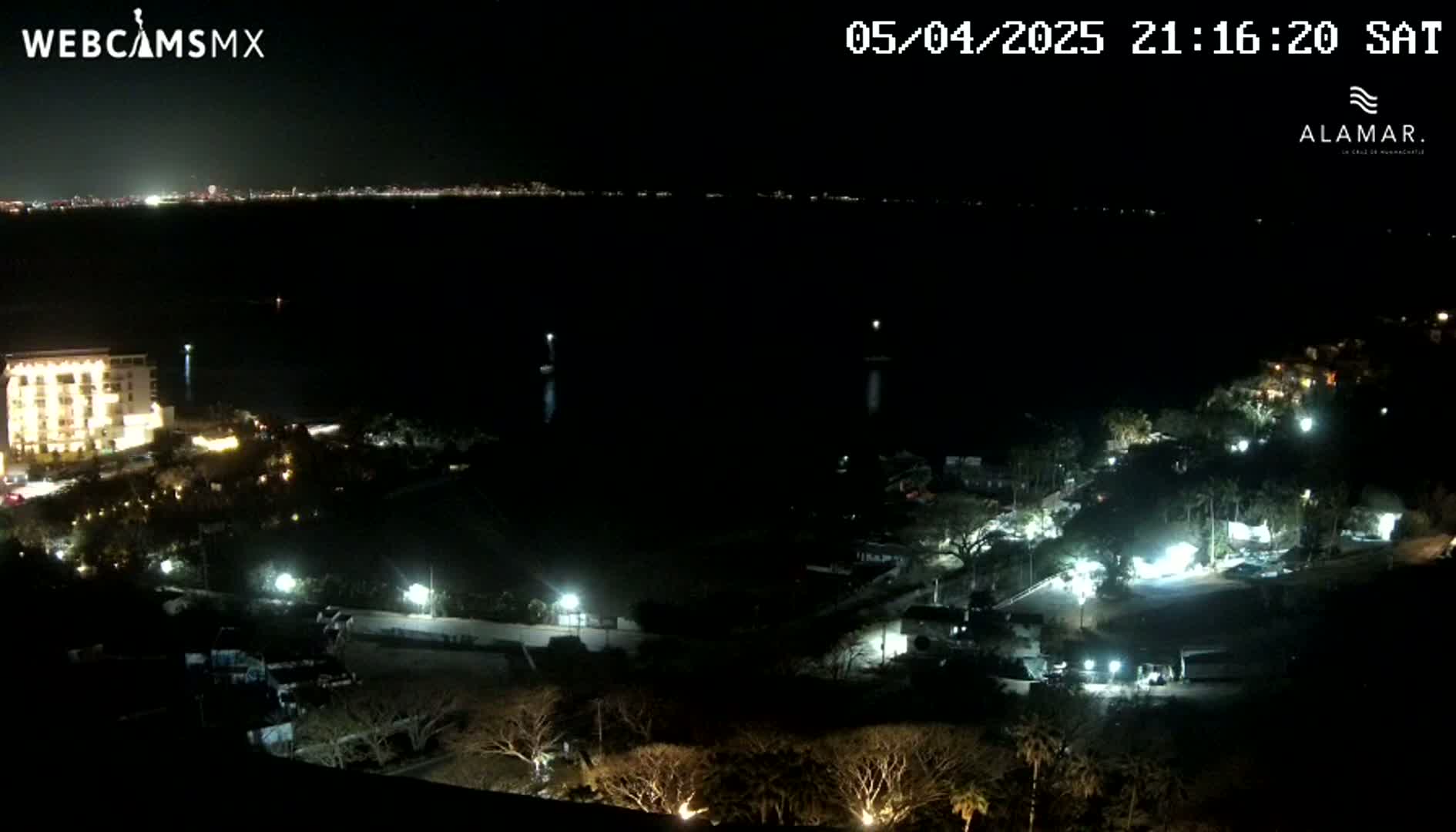 A nighttime, high-angle view shows a coastal area with a lit-up building, scattered lights, and dark trees, overlooking a dark body of water with a distant city skyline visible on the horizon.