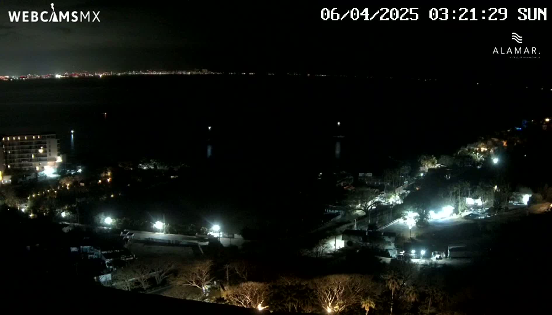 A nighttime, high-angle view shows a dark coastline with sparsely lit buildings and trees, overlooking a dark body of water with a distant, faintly lit city skyline.
