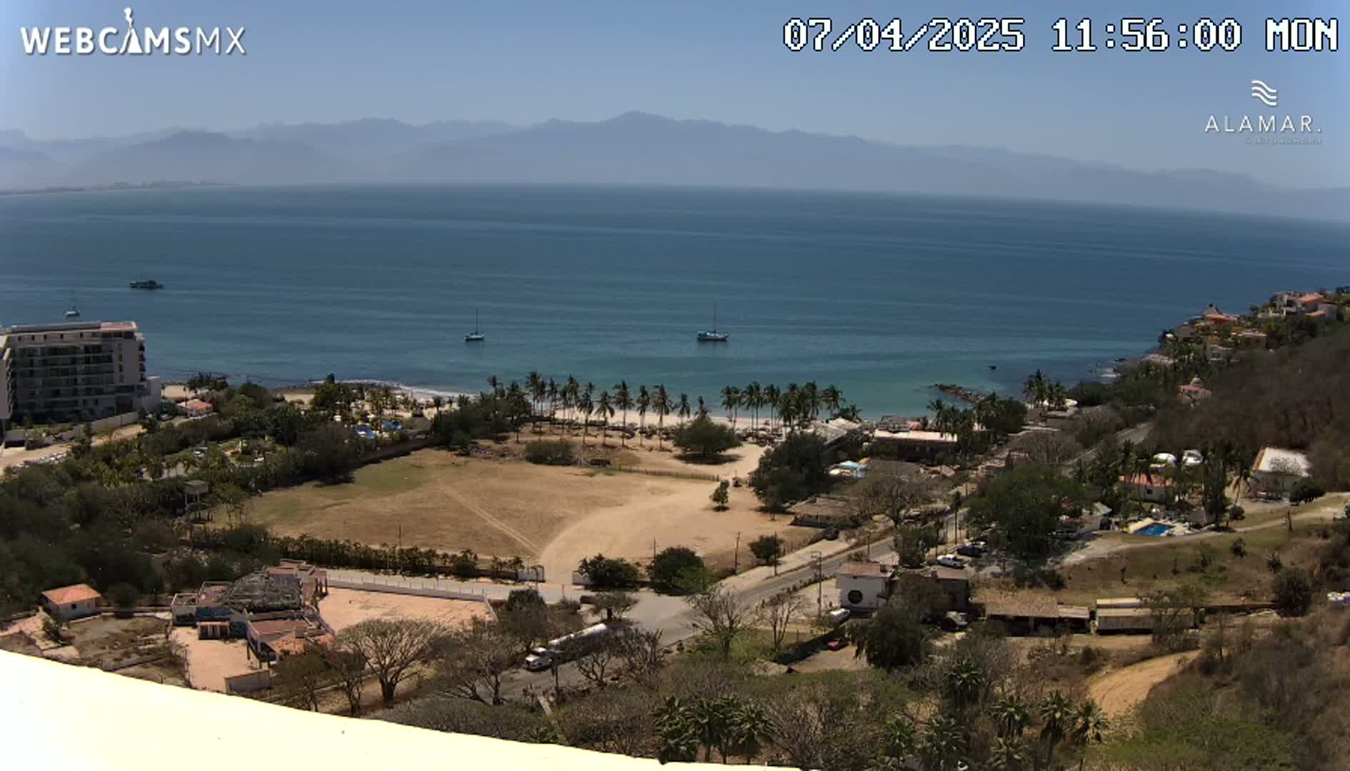 A coastal town scene on a clear, sunny day, showing a sandy beach lined with palm trees, a calm ocean with a few boats, and a hilly landscape with buildings and vegetation.