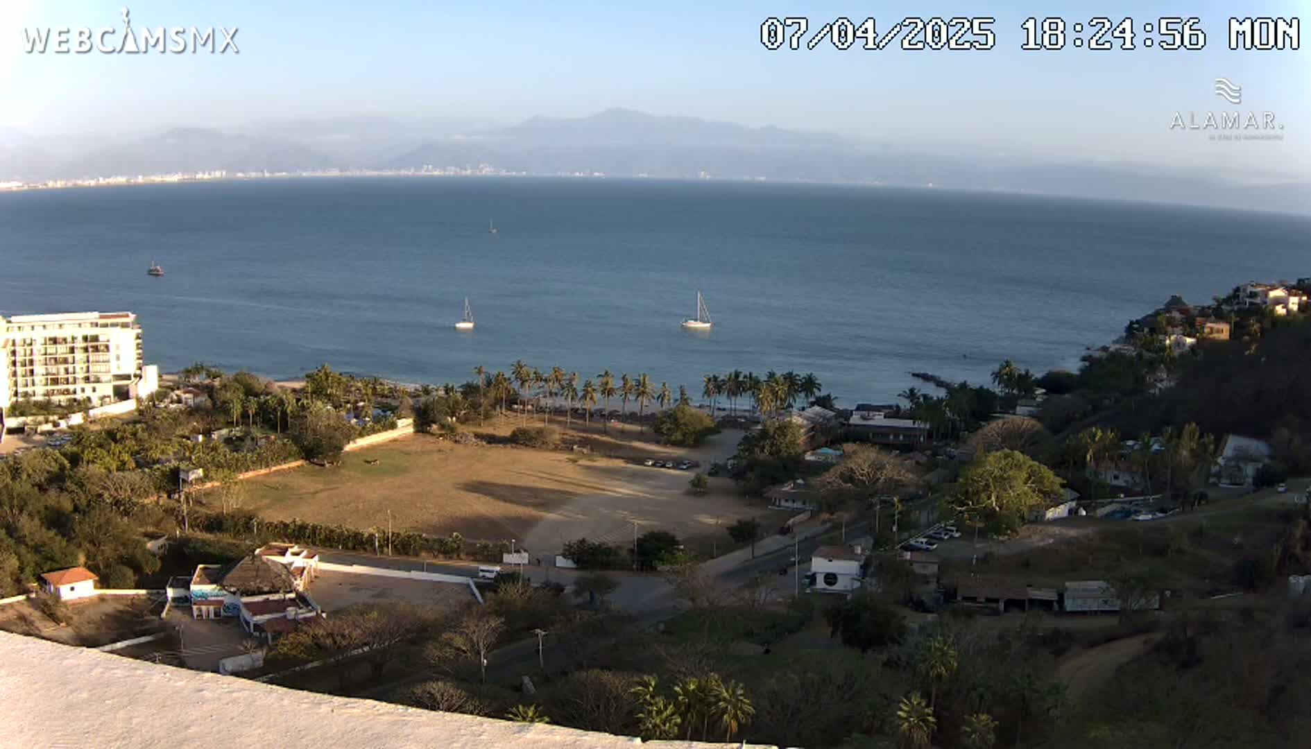 An aerial view of a coastal town at sunset, showing a calm ocean with a few sailboats, a beach with palm trees, buildings, and a large, empty field.