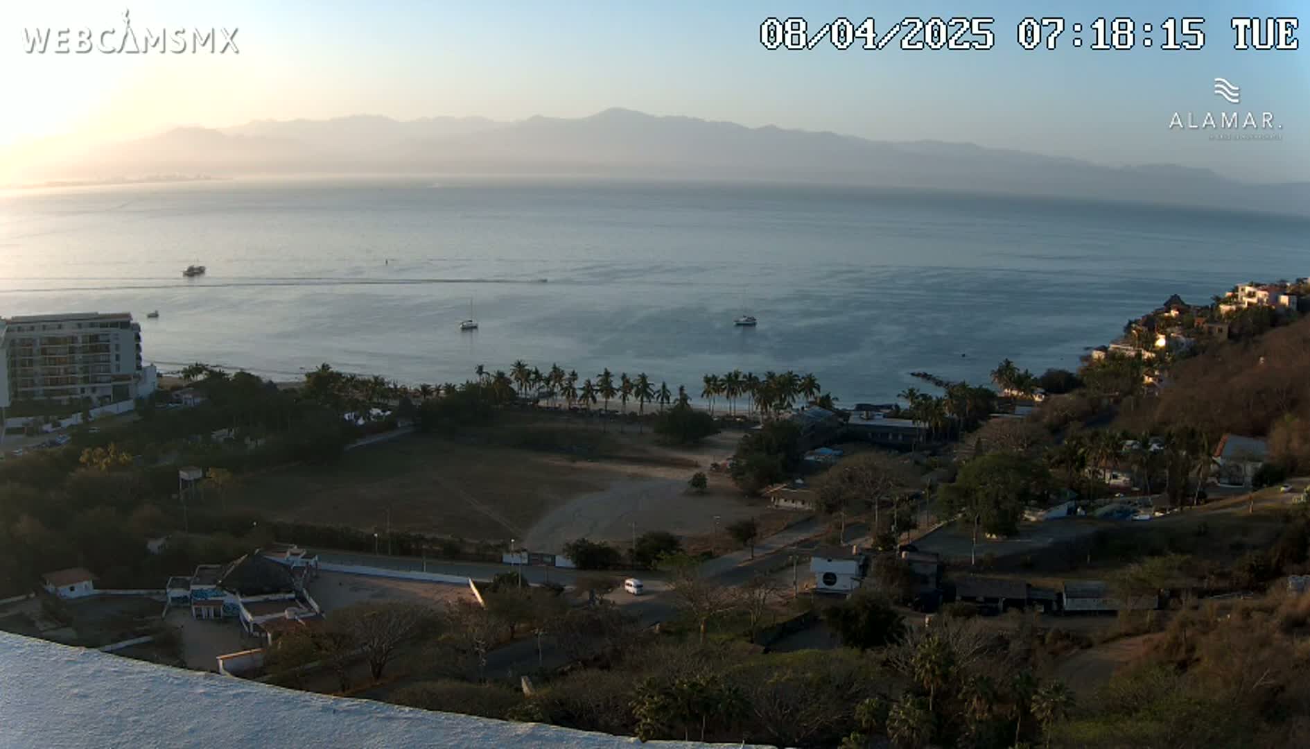 A high-angle, long shot of a coastal scene at sunrise, showing a calm ocean, a sandy beach lined with palm trees, buildings, and hazy mountains in the distance.