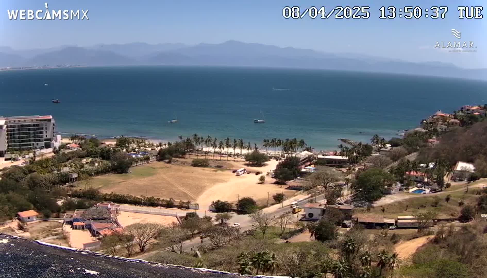 An elevated view shows a beach with palm trees, a calm ocean, mountains in the distance, and coastal buildings under sunny skies.