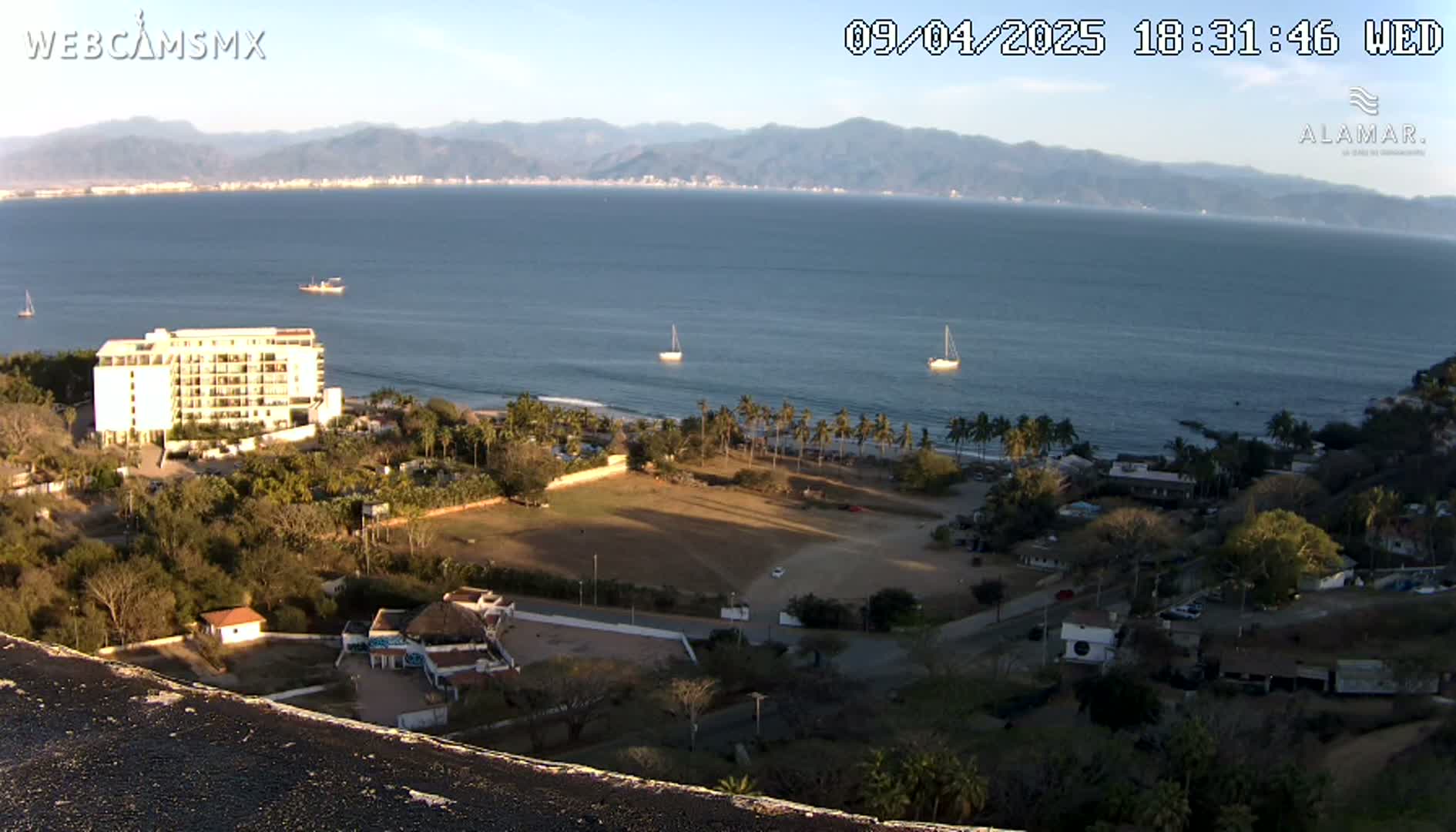A high-angle view of a coastal scene at sunset, showing a large hotel, palm trees, a sandy beach, calm ocean water with sailboats, and distant mountains under a clear sky.