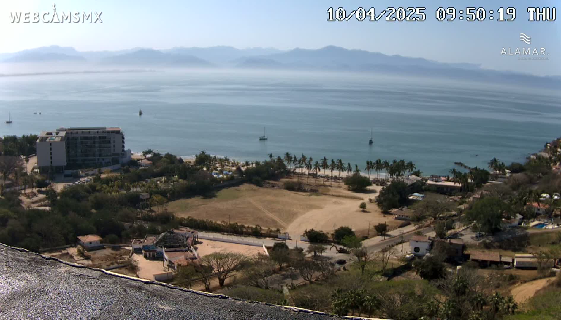 An elevated view shows a calm ocean with a few boats, a sandy beach lined with palm trees, coastal buildings, and hazy mountains in the distance under a clear sky.