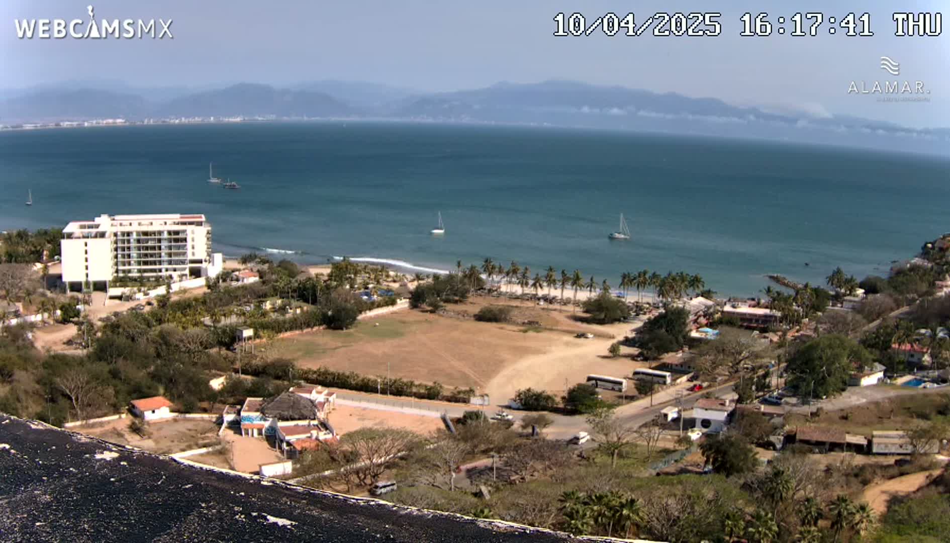 A high-angle, sunny view shows a coastal town with a beach, ocean, and a large building, all situated against a backdrop of distant mountains.