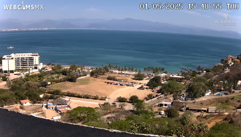 A coastal scene on a sunny day shows a large body of water, a sandy beach with palm trees, a hotel, and various buildings and parking lots nestled amongst green vegetation.