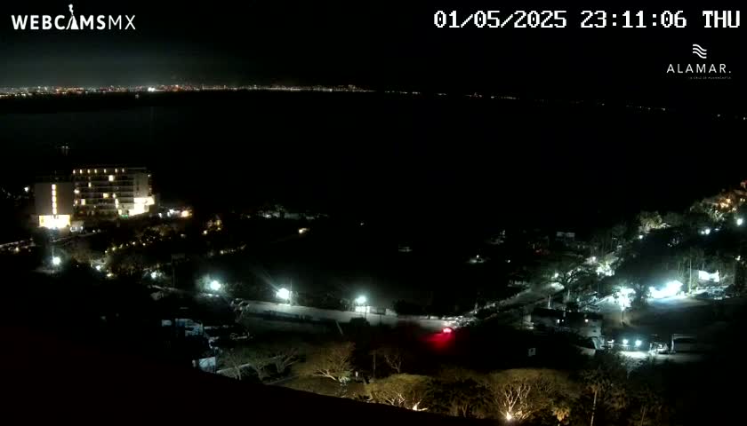 A nighttime view of a coastal area shows a lit-up hotel and other buildings near a dark body of water, under a clear sky.