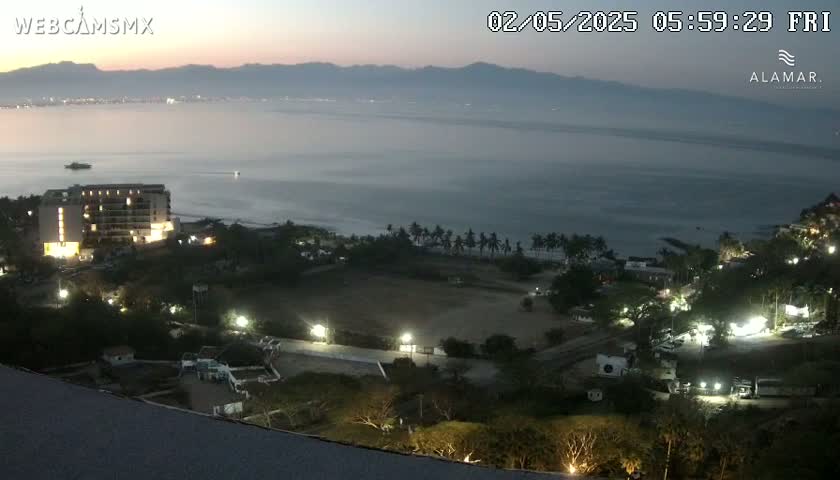 A coastal scene at twilight shows a calm body of water, a hotel, and a sparsely lit area of land with palm trees, under a clear, dark sky.