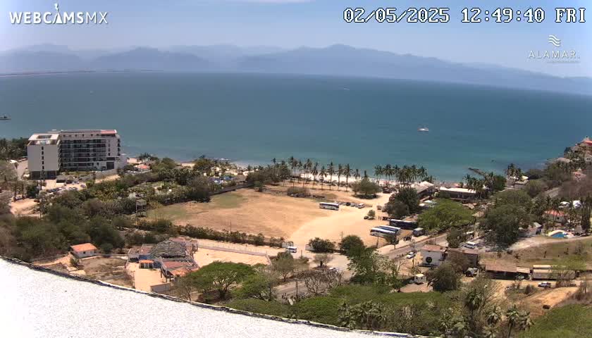 An aerial view shows a beach with palm trees, a hotel, and other buildings on a sunny day, overlooking a calm ocean and distant mountains.