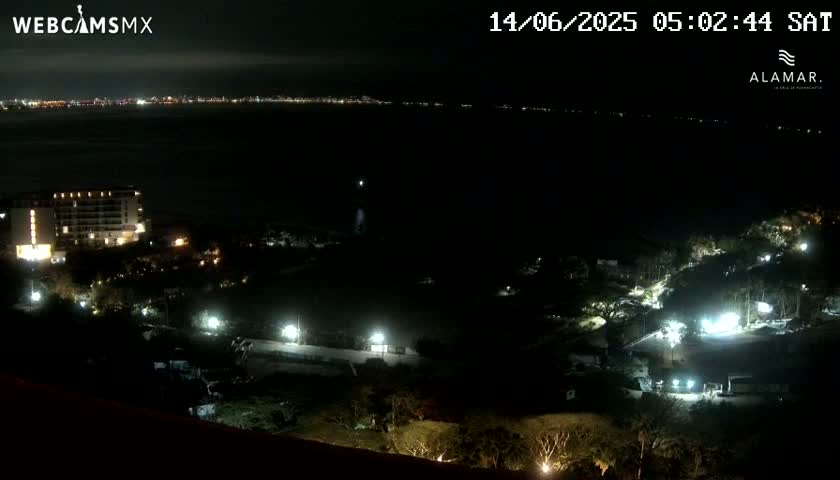 A nighttime view shows a coastal area with a large building and surrounding lights, overlooking a dark body of water with a distant city skyline illuminated in the background under a clear sky.