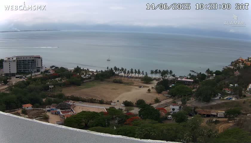 A high-angle view of a coastal scene under overcast skies shows a hotel, a beach with palm trees, and a large, empty field, all overlooking a calm ocean.