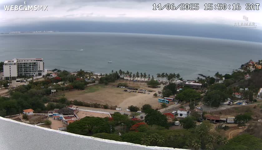 A high-angle view of a coastal scene under overcast skies shows a hotel, a beach with palm trees, and a large, empty field, all overlooking a calm ocean.