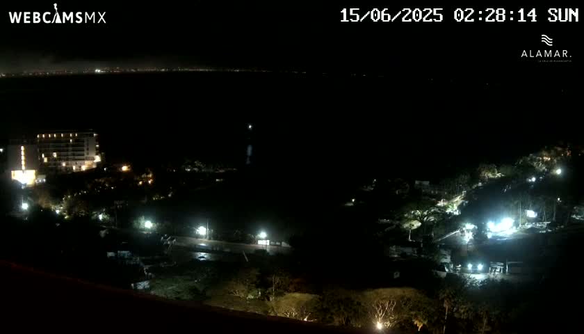 A nighttime view shows a coastal area with a multi-story building and sparsely lit vegetation sloping down to a dark body of water with a faint city glow on the horizon.