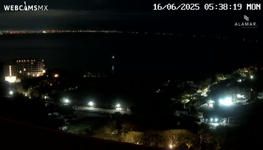 A nighttime view of a coastal area shows a city skyline across a dark body of water, with a lit-up building and other lights scattered across a hillside below.