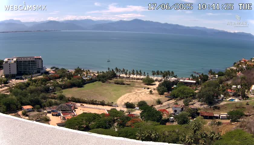 An aerial view shows a coastal scene on a sunny day, featuring a large body of water, a beach lined with palm trees, a hotel, and various buildings nestled among lush green vegetation, all backed by distant mountains.