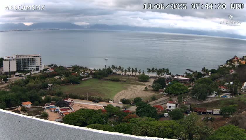 An aerial view shows a coastal town on a cloudy day, with a calm ocean stretching to distant mountains, palm trees lining the beach, and a large building visible near the water.