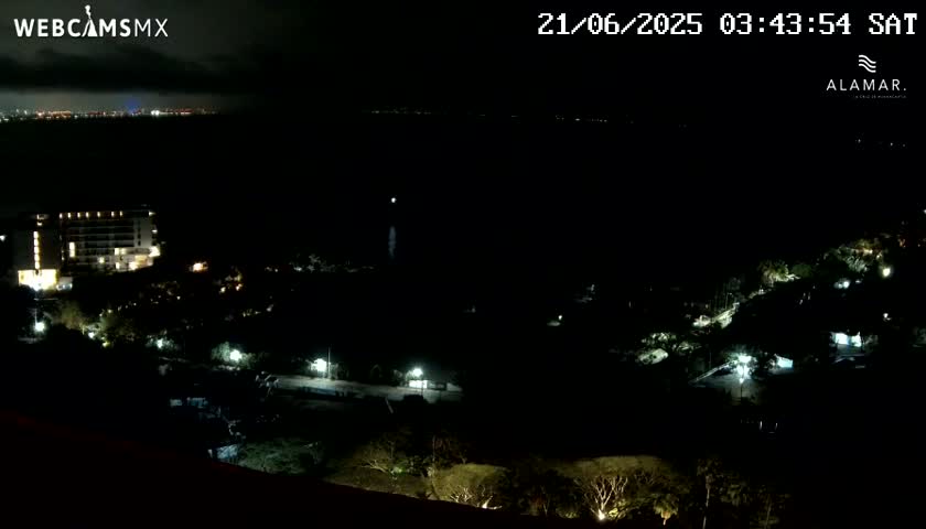A nighttime view of a coastal area shows a lit-up hotel and surrounding vegetation under a partly cloudy sky.