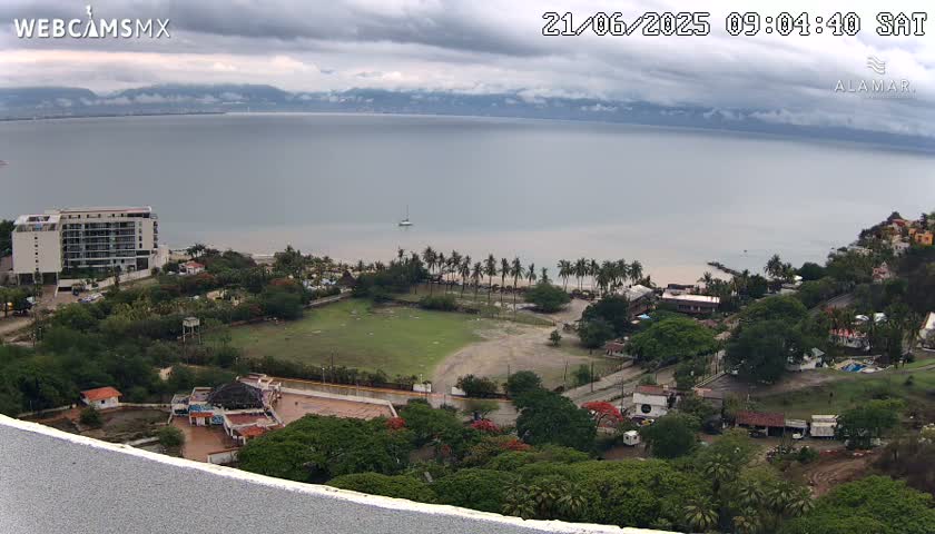 A coastal scene under cloudy skies shows a large body of water, a beach with palm trees, a grassy field, buildings, and lush green vegetation.