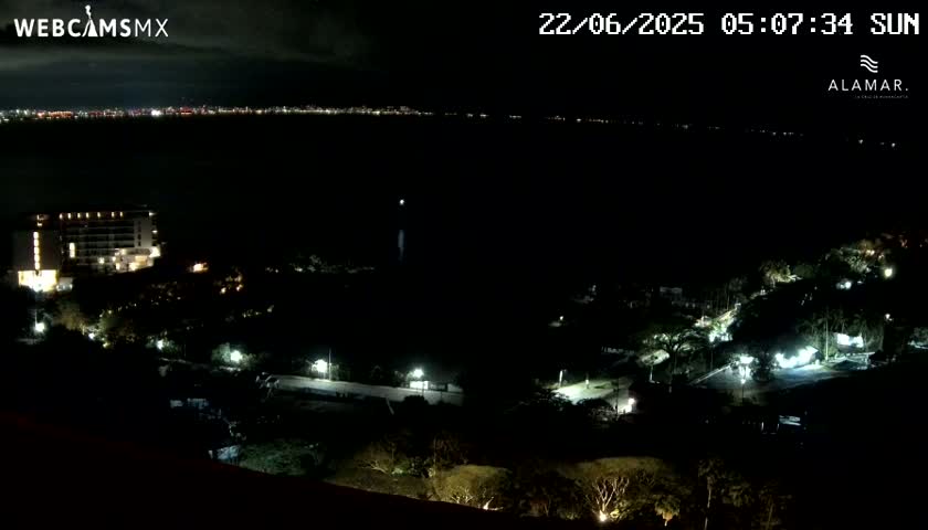 A nighttime view of a coastal area shows a lit-up hotel and other buildings near a dark body of water with a distant city skyline visible across the water.