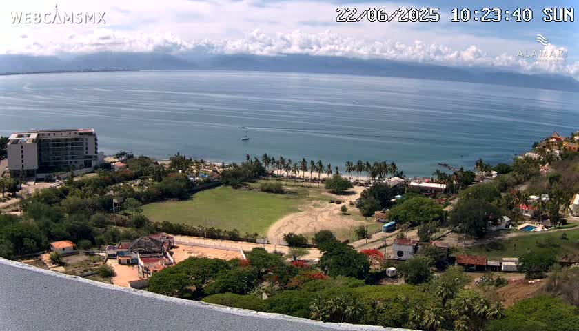 A coastal scene under partly cloudy skies shows a beach with palm trees, a large building, and other structures nestled among lush green vegetation, all overlooking a calm ocean and distant mountains.