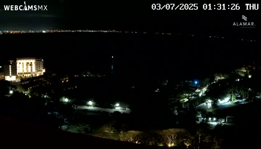 A nighttime aerial view shows a brightly lit hotel and surrounding landscape, with a distant city skyline visible across a dark body of water under a clear sky.