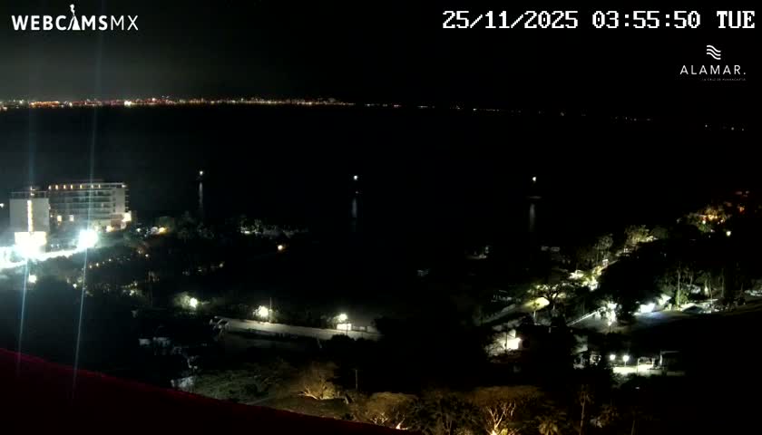 The image captures a clear nighttime view of a coastal area, featuring a brightly lit building and illuminated landscaping in the foreground, with a distant city skyline across a dark body of water.