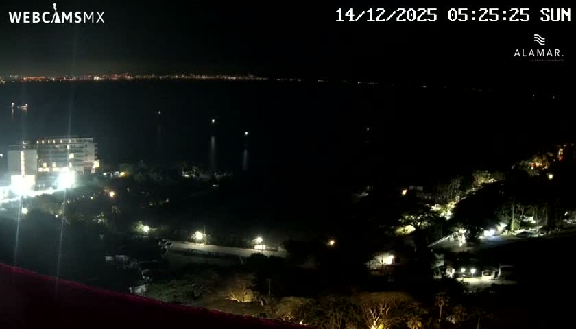 The image captures a clear nighttime view of a coastal area, featuring a brightly lit building and illuminated landscaping in the foreground, with a distant city skyline across a dark body of water.
