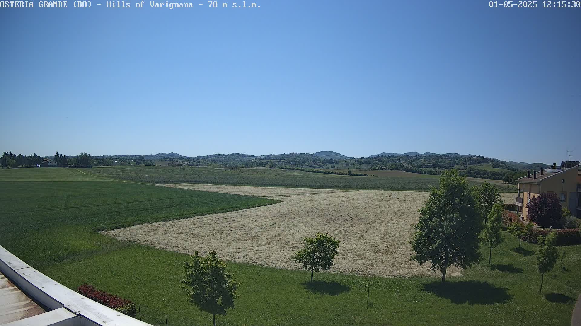 Under a clear blue sky, a sprawling landscape of green fields and recently harvested hay stretches towards rolling hills in the distance.