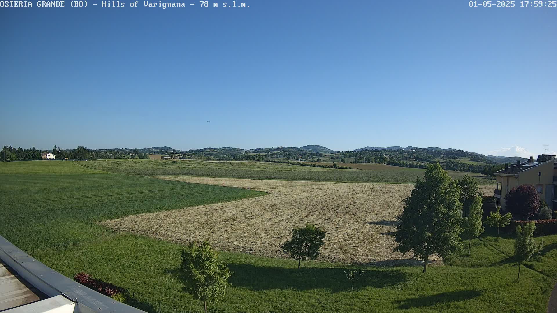 A clear blue sky overlooks a vast expanse of farmland, with patches of harvested hay and growing crops, punctuated by a few trees and a distant building under a bright sun.