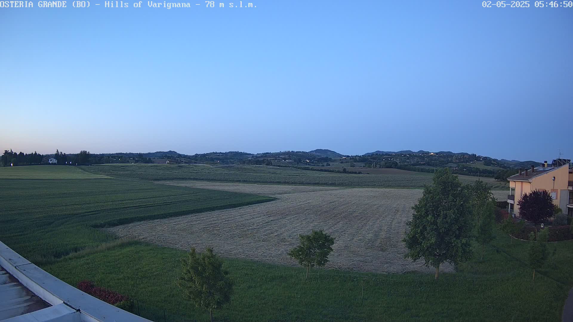 Under a clear, twilight sky, a flat expanse of farmland with harvested and unharvested fields stretches toward rolling hills in the distance, punctuated by a few buildings and trees.