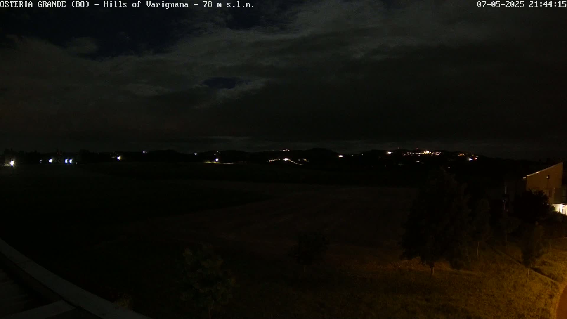 A nighttime view of dark hills and a flat field, with a few distant lights visible under a cloudy sky.
