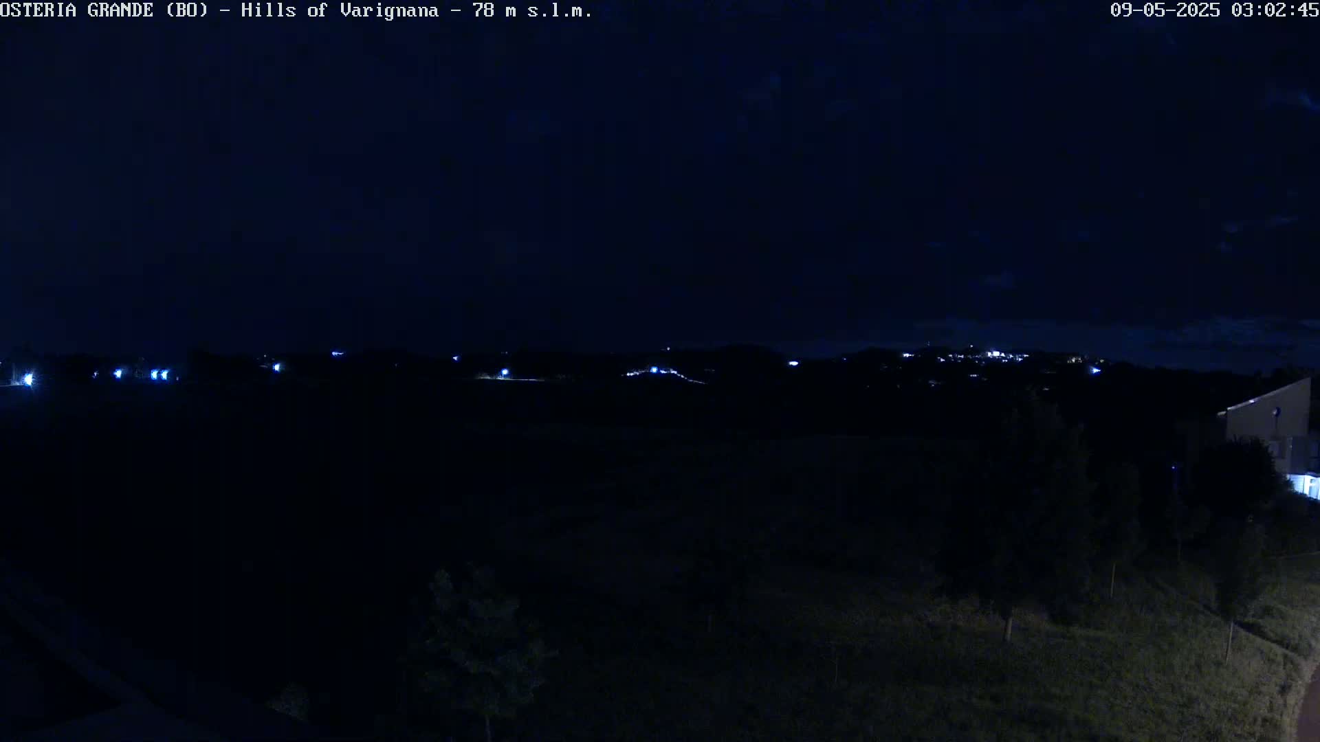 A nighttime view of dark hills dotted with a few faint lights, under a mostly clear, dark sky.