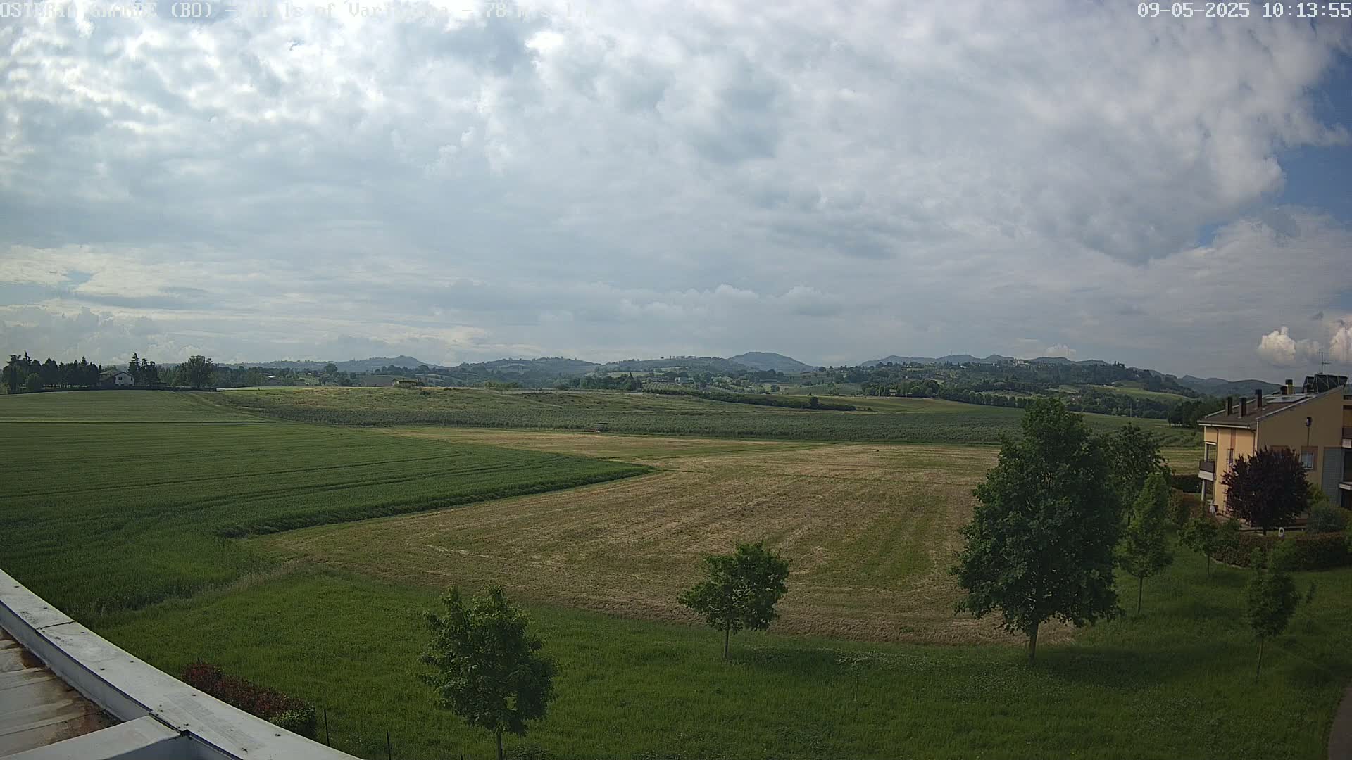 A partly cloudy sky overlooks a vast expanse of green and brown farmland, punctuated by a few trees and a building in the lower right corner.