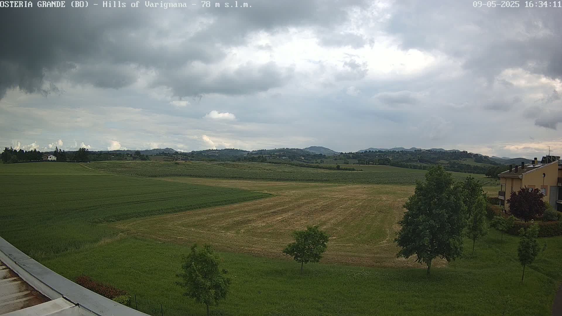 A partly cloudy day reveals expansive green fields, distant rolling hills, and a few buildings in the foreground.