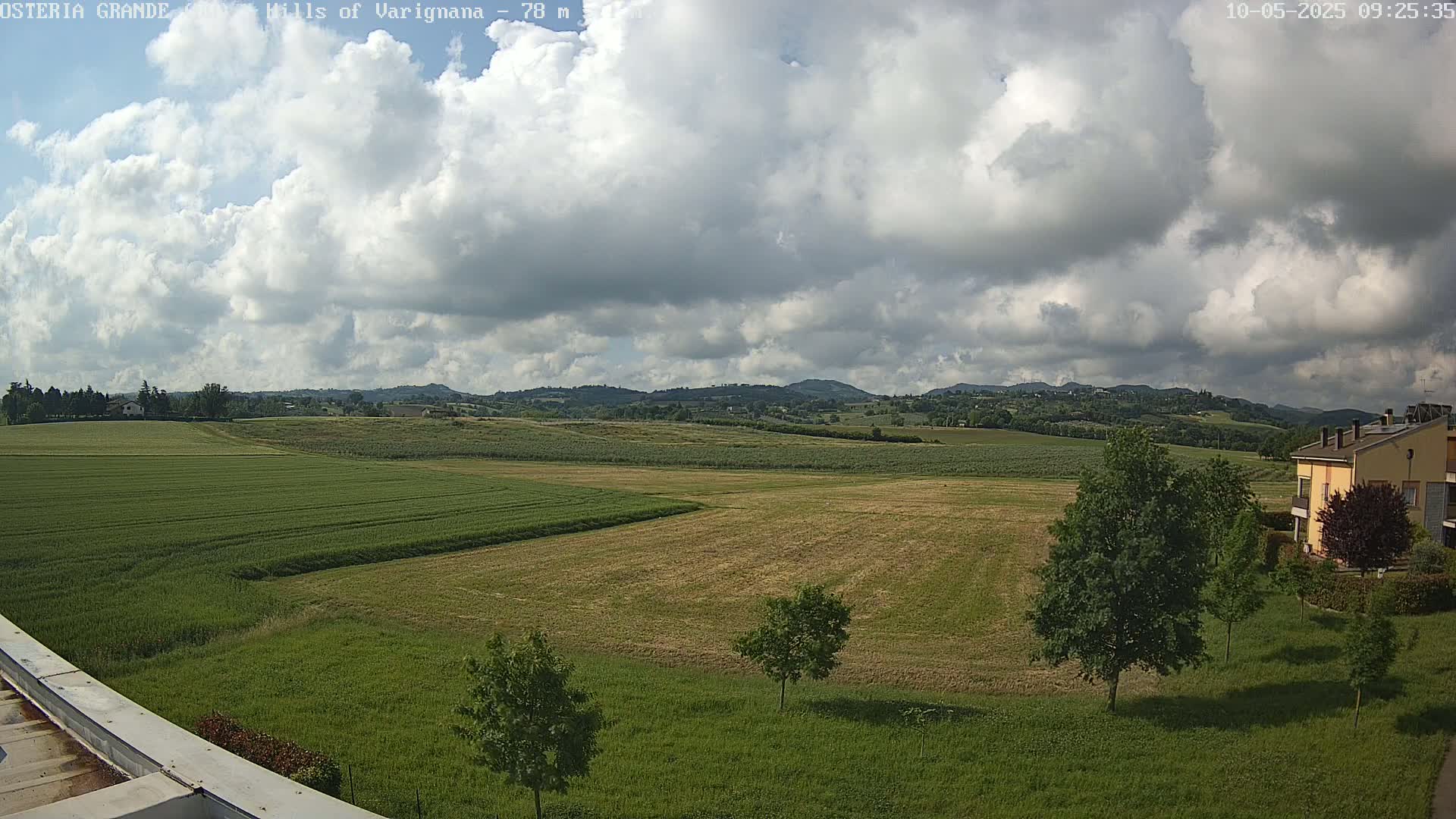 A partly cloudy sky overlooks expansive fields of varying shades of green and tan, dotted with a few trees and a building in the distance.