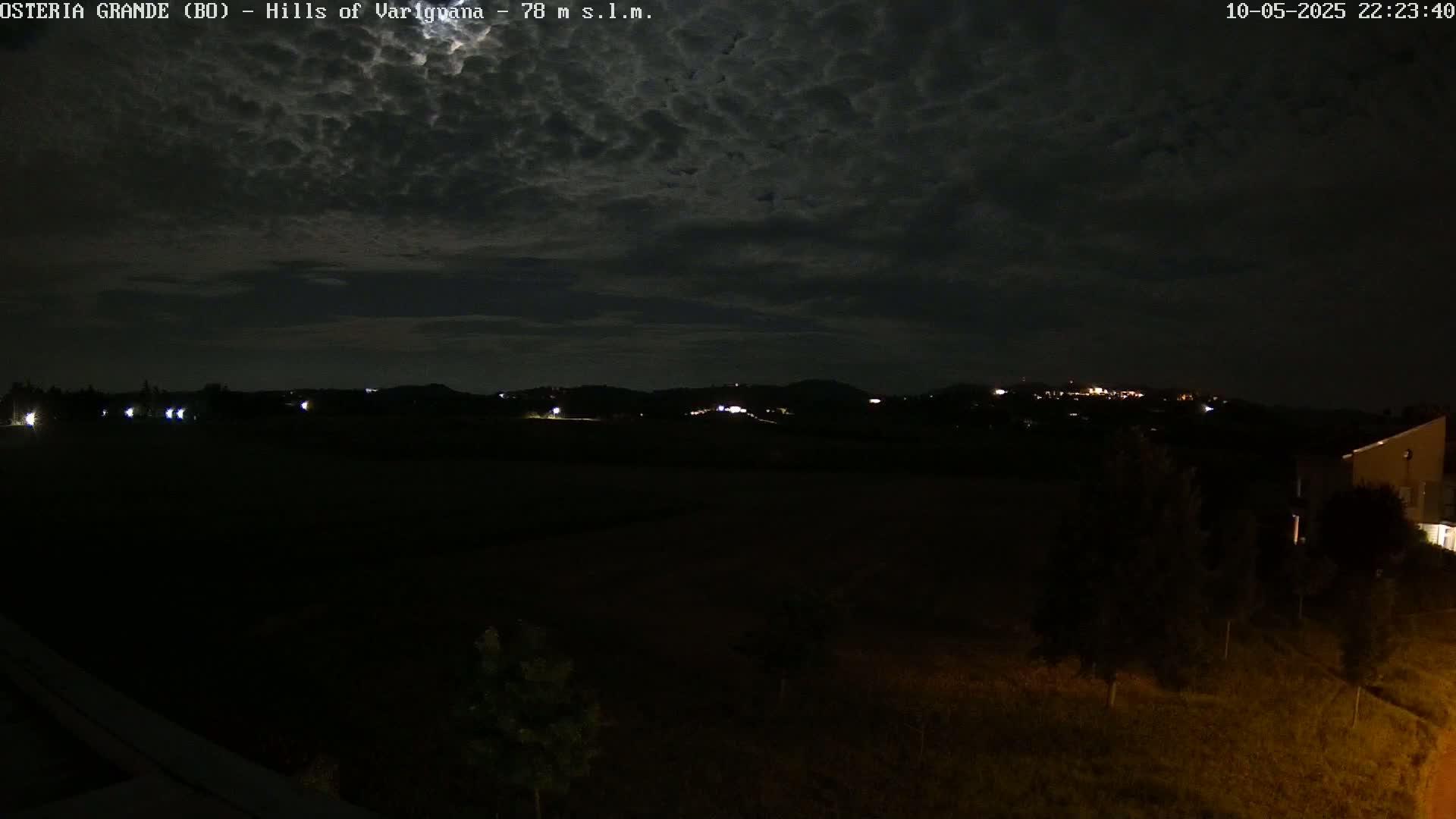 A nighttime view of dark, rolling hills under a cloudy sky, illuminated by a few distant lights.