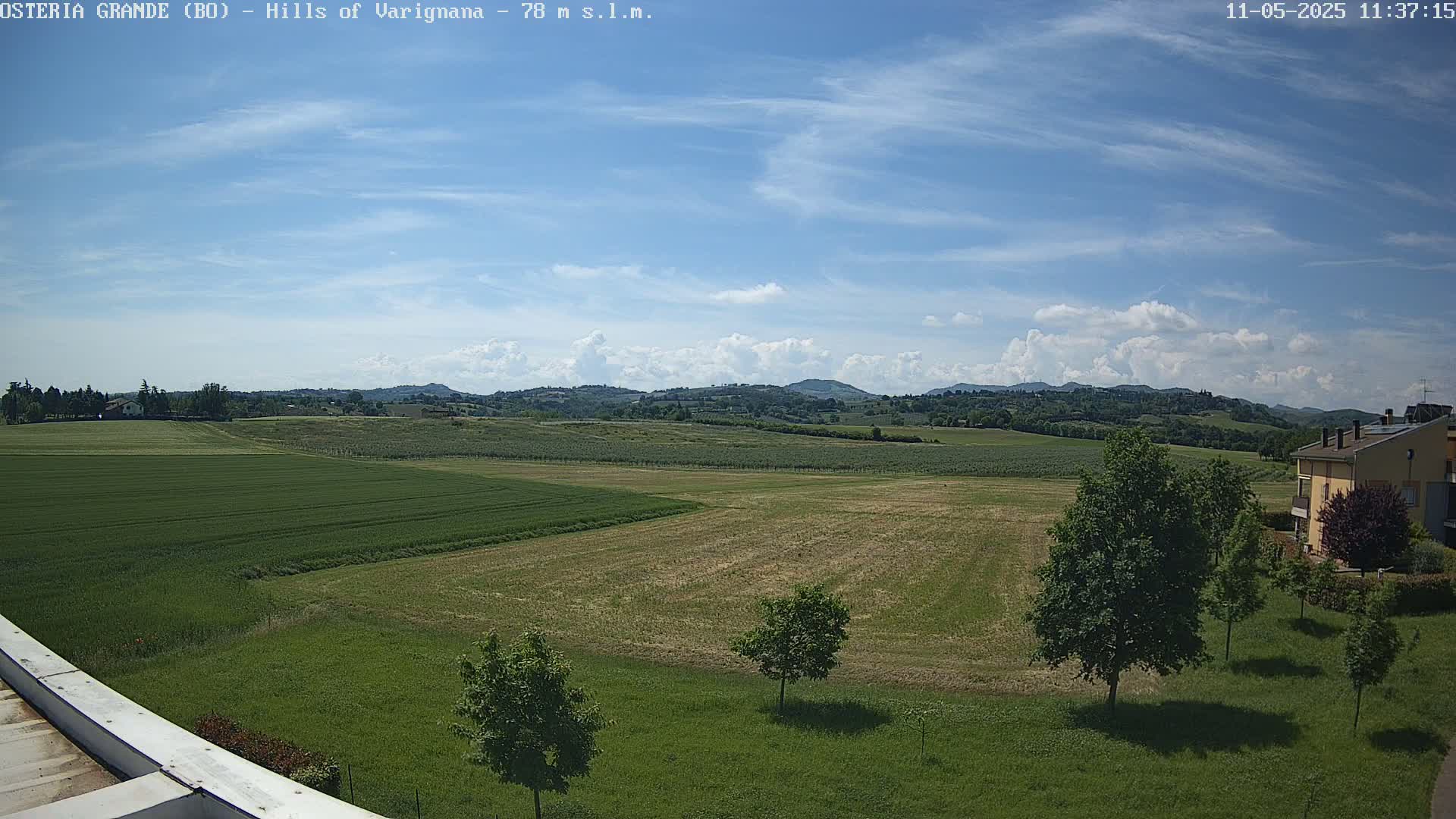 A partly cloudy sky overlooks expansive green fields and distant rolling hills, with a few trees and a building visible in the foreground.
