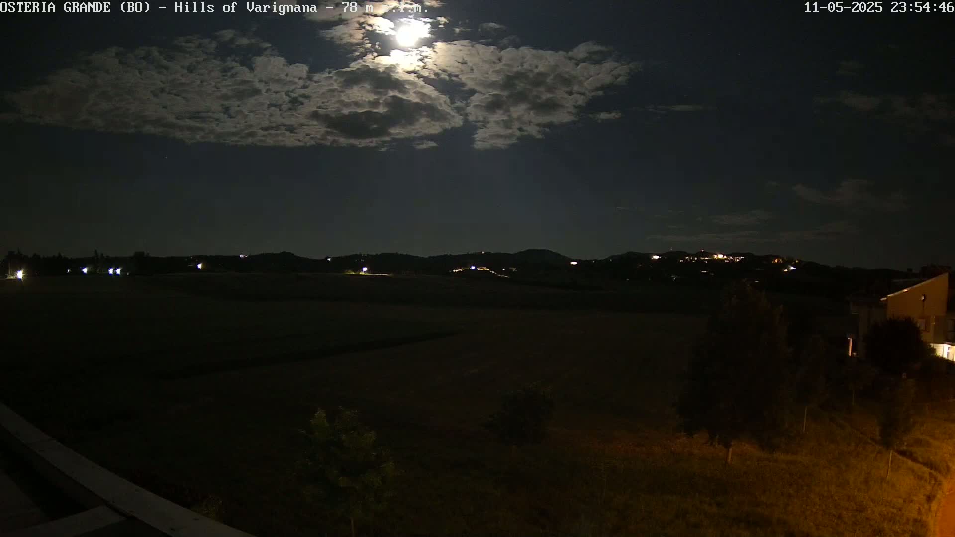 A nighttime view of dark, rolling hills under a partly cloudy sky illuminated by a bright moon.
