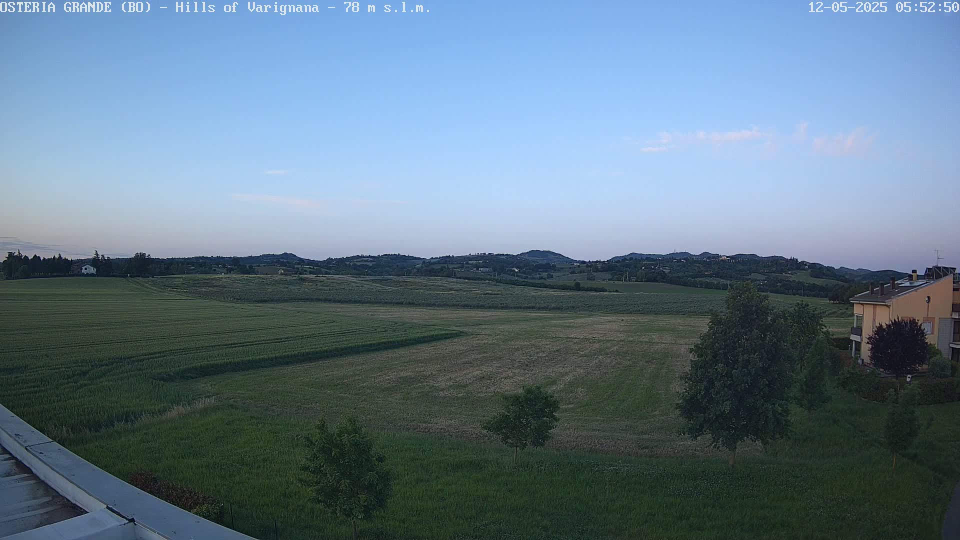 Under a clear, twilight sky, a vast expanse of verdant farmland stretches towards rolling hills in the distance, with a few buildings visible near the bottom right.