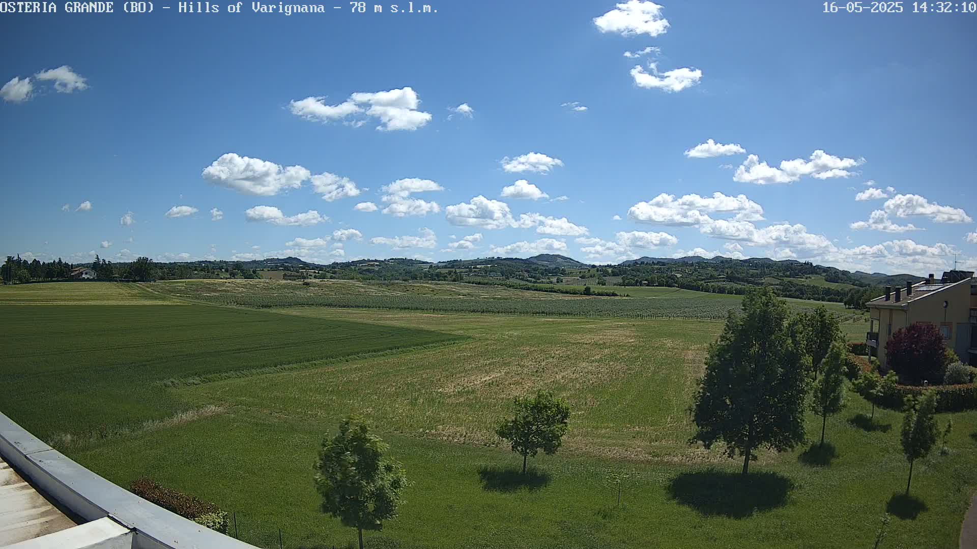 A sunny day with scattered cumulus clouds over a vast green field, dotted with trees and distant rolling hills.