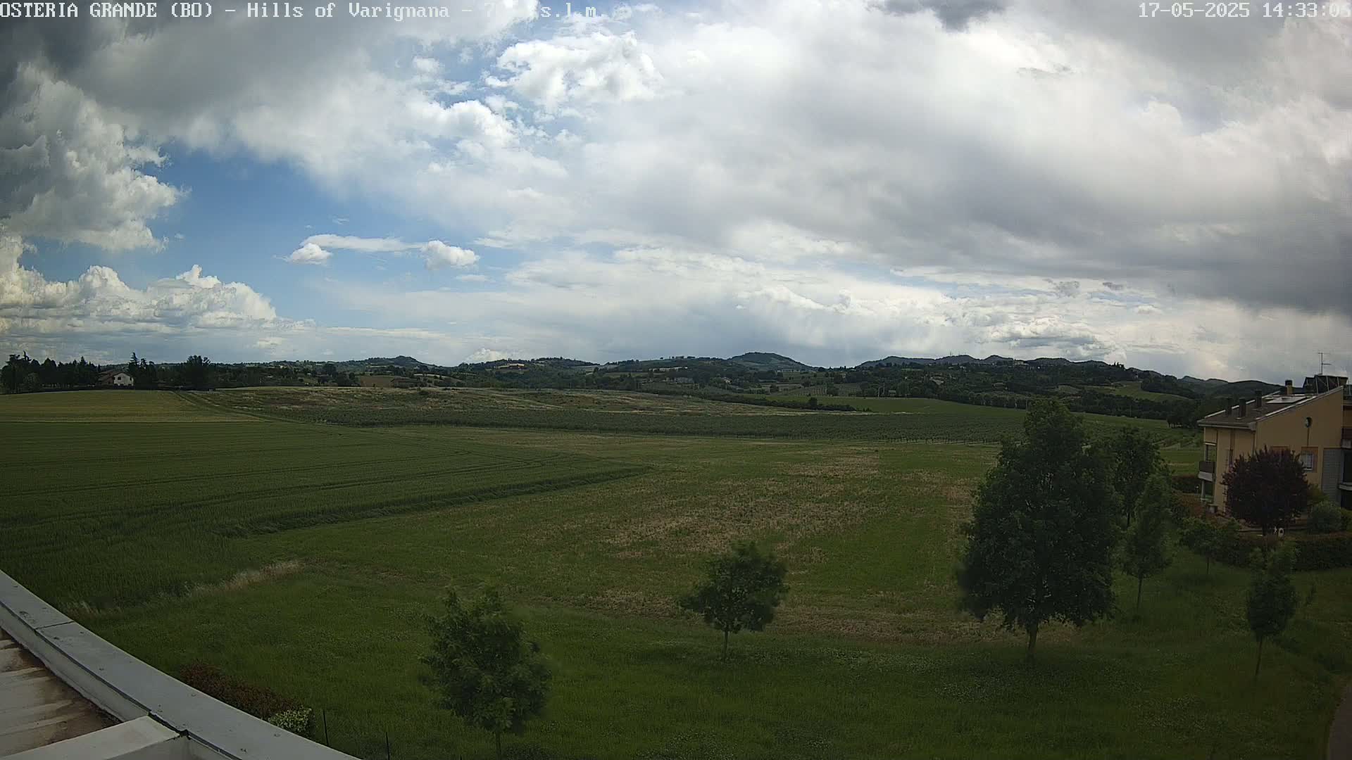 A partly cloudy sky overlooks a vast, green field dotted with trees, leading to rolling hills in the distance.