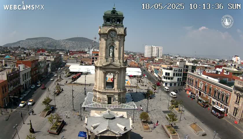 A city square with a tall clock tower at its center, surrounded by low buildings, under a clear, sunny sky.