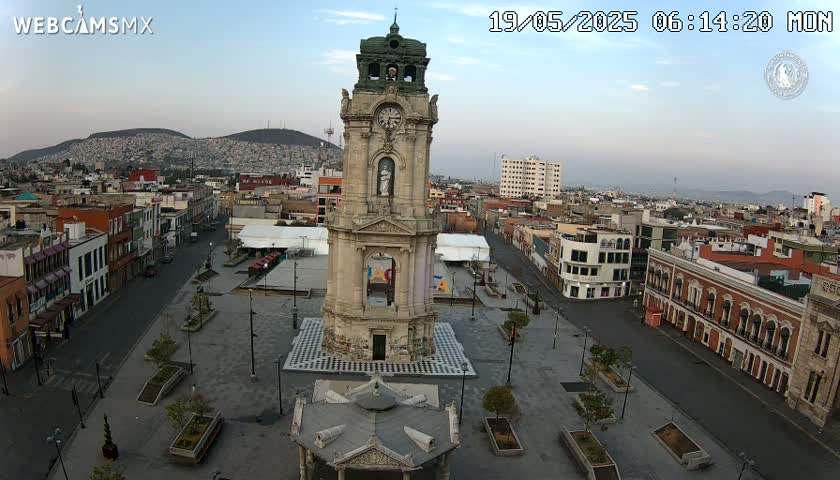 A tall clock tower stands in a mostly empty plaza in a city under a clear morning sky, with mountains visible in the distance.