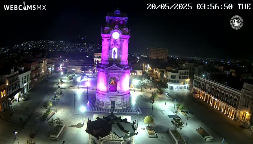 An illuminated clock tower stands in a city square at night, surrounded by buildings and landscaping under clear skies.