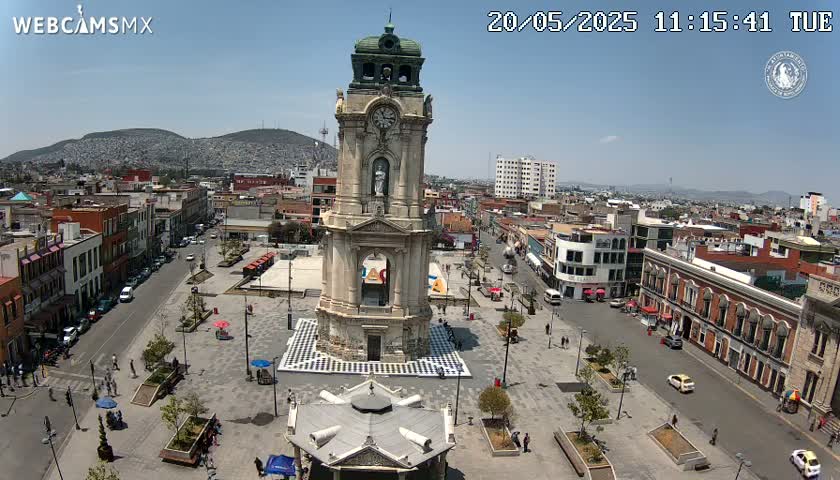 A high-angle view of a town square on a sunny day, centered on a tall clock tower surrounded by buildings and a paved plaza with sparse pedestrian traffic.