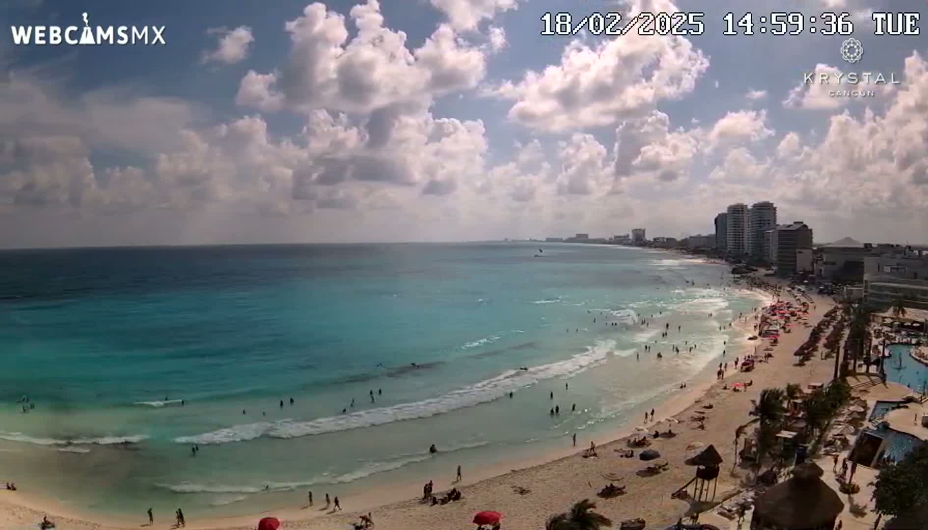 A partly cloudy day shows a beach with light waves, many people swimming and sunbathing, and high-rise buildings in the distance.