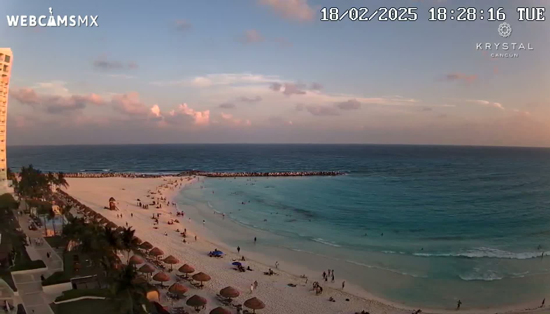 A beach with turquoise water and a sandy shore, populated with people and beach umbrellas, is seen under a partly cloudy sunset sky.