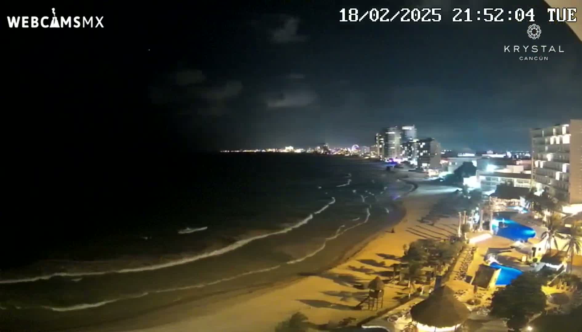 A nighttime beach scene shows gently lapping waves on a sandy shore, with illuminated hotels and city lights visible in the distance under a partly cloudy sky.