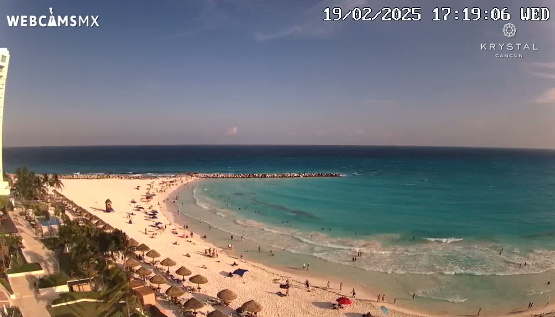 A beach with turquoise water and a sandy shore is populated with bathers under a clear sky.