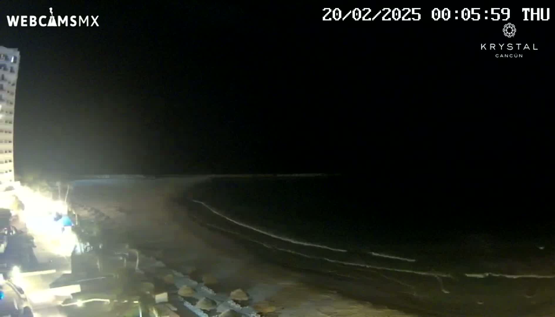 A nighttime view of a dark beach with gentle waves lapping the shore, illuminated by lights from buildings and vehicles along the beachfront.